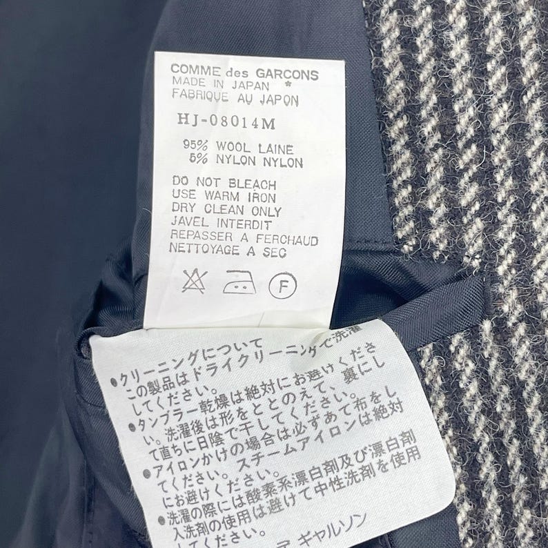 May include: A black and white checkered fabric swatch with a white tag attached. The tag has black text that reads "COMME des GARCONS MADE IN JAPAN FABRIQUE AU JAPON HJ-08014M 95% WOOL LAINE 5% NYLON NYLON DO NOT BLEACH USE WARM IRON DRY CLEAN ONLY JAVEL INTERDIT REPASSER A FERCHAUD NETTOYAGE A SEC" and includes a washing instruction symbol.