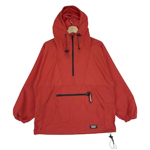Könnte beinhalten: Rote Anorak-Jacke mit Kapuze und Reißverschluss. Die Jacke hat eine Vordertasche mit schwarzem Reißverschluss und einen kleinen Logoaufnäher. Die Kapuze hat Kordelzüge. Die Jacke hängt an einem Holzkleiderbügel.