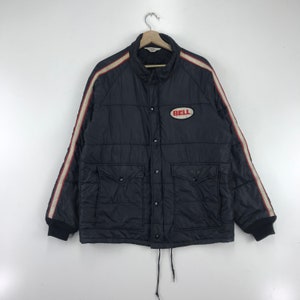 May include: A black quilted jacket with a white and red stripe down the sleeves. The jacket features two front pockets and a "BELL" patch on the chest. The jacket is suspended from a hanger.