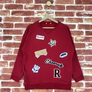 Peut inclure: Sweat-shirt rouge avec divers patchs brodés colorés, dont "PandaZ", "Cheer Up R", "Strategy", "Vespa" et une étoile.