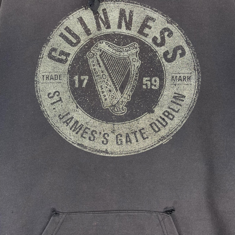 May include: Dark grey hoodie with a distressed circular graphic. The graphic features the word "GUINNESS" and "ST. JAMES'S GATE DUBLIN", along with a harp and the numbers "17" and "59".
