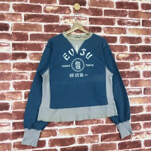 May include: A blue and grey sweatshirt with the word "EVISU" in a white circle on the front. The circle also has the words "OSAKA" and "TOKYO" on either side of it. Below the circle is a white circle with Japanese characters and the words "初 第 一".