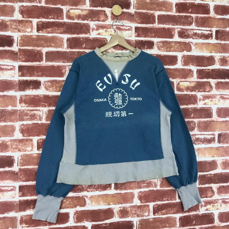 May include: A blue and grey sweatshirt with the word "EVISU" in a white circle on the front. The circle also has the words "OSAKA" and "TOKYO" on either side of it. Below the circle is a white circle with Japanese characters and the words "初 第 一".