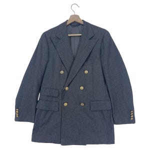 Puede incluir: Un blazer gris de doble botonadura con botones dorados. El blazer tiene dos filas de botones, solapas con muescas y dos bolsillos delanteros. Las mangas tienen botones en los puños. La chaqueta está colgada de una percha de madera.