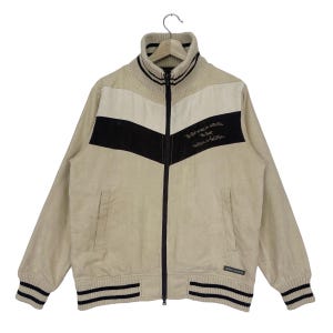 May include: A beige corduroy jacket with a black and white color block design. The jacket has a zipper closure and a high collar. The text "The best way to settle the debt is to forget it" is embroidered on the jacket.