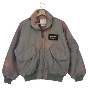 以下が含まれることがあります： ジッパー留めと「AVIATOR U.S.A.F.」と書かれたパッチが付いた緑色のボンバージャケット