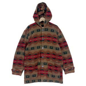 Op de afbeelding: Een bruine capuchontrui met een kleurrijk geometrisch patroon. De trui heeft een knoopsluiting aan de voorkant en twee zakken.