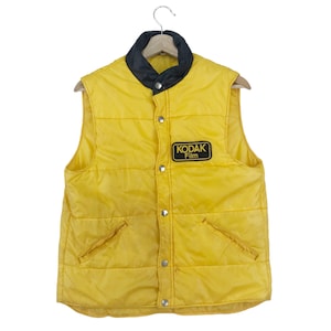 May include: A yellow quilted vest with a black collar and a snap closure. The vest has two pockets on the front and a patch on the left side that says "Kodak Film".
