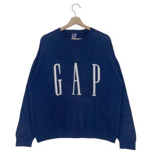 May include: A navy blue GAP sweatshirt with the brand name in large, white block letters. The sweatshirt has a crew neck and long sleeves. A smaller GAP logo is visible near the neckline.