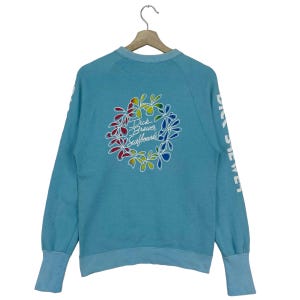 Puede incluir: Una sudadera azul claro con un diseño de flores de colores y el texto "Dick Brewer Surfboards" en la espalda. Las mangas tienen el texto "Dick Brewer" impreso.