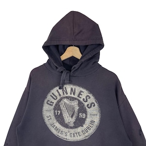 May include: Dark grey hooded sweatshirt featuring the Guinness logo. The logo is a white circular design with the word "Guinness" and a harp. The text "St. James's Gate Dublin" is also visible.