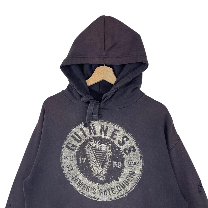 May include: Dark grey hooded sweatshirt featuring the Guinness logo. The logo is a white circular design with the word "Guinness" and a harp. The text "St. James's Gate Dublin" is also visible.