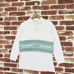 Op de afbeelding: Een lichtroze sweatshirt met lange mouwen en een grijze horizontale streep over de borst. De tekst "courr&egrave;ges paris" staat boven de streep en "courr&egrave;ges sport futur" staat op de streep. Het sweatshirt hangt aan een houten hanger.