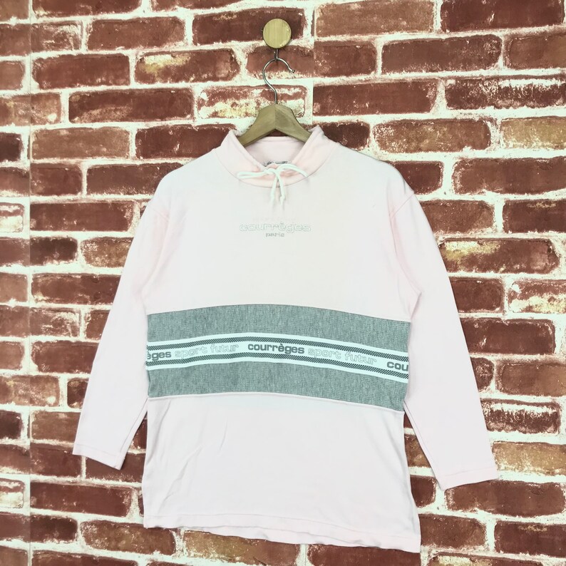 Op de afbeelding: Een lichtroze sweatshirt met lange mouwen en een grijze horizontale streep over de borst. De tekst "courr&egrave;ges paris" staat boven de streep en "courr&egrave;ges sport futur" staat op de streep. Het sweatshirt hangt aan een houten hanger.
