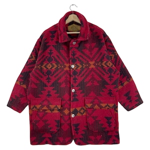 Op de afbeelding: Een rood en zwart patroonjas met een knoopsluiting, twee voorzakken en een kraag. De jas heeft een geometrisch ontwerp in tinten rood, zwart en oranje. De jas hangt aan een houten hanger.