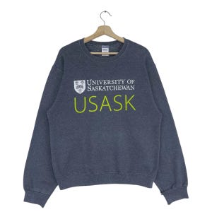 May include: A dark grey sweatshirt featuring the text "UNIVERSITY OF SASKATCHEWAN" and "USASK" in white and lime green. The sweatshirt is displayed on a wooden hanger.