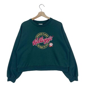 Può includere: Una felpa verde scuro con collo rotondo bianco e maniche lunghe. La parte anteriore della felpa presenta un logo circolare con il testo "Frosted Flakes Kellogg's The Best To You" in lettere rosse e gialle. Un personaggio dei cartoni animati è raffigurato nel logo.