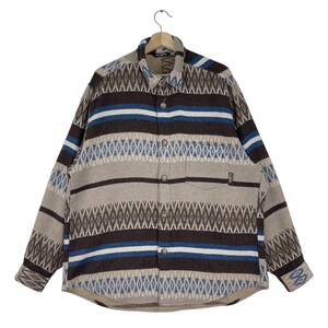 Op de afbeelding: Een overhemd met knopen en een geometrisch patroon in bruin, blauw en beige tinten. Het overhemd heeft lange mouwen, een borstzak en een kraag. Het hangt aan een houten kleerhanger.