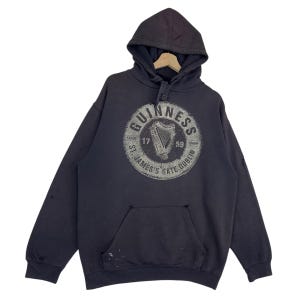 May include: Dark grey hooded sweatshirt with a front pocket and a distressed Guinness logo. The logo features a harp and the text "Guinness St. James's Gate Dublin". The sweatshirt has a drawstring hood and long sleeves.