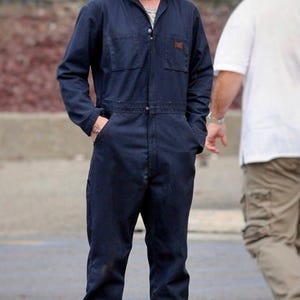 May include: A man wearing a dark blue jumpsuit with a zipper and pockets. He is wearing black work boots and has short blond hair.
