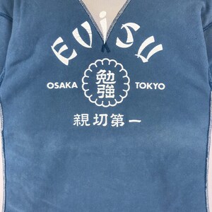 May include: A blue long-sleeved shirt with a white graphic design. The graphic features the text "EVISU" in a curved arc, with the words "OSAKA" and "TOKYO" on either side. A circular graphic with Japanese characters is centred below the text. The text "最初第一" is below the circular graphic.