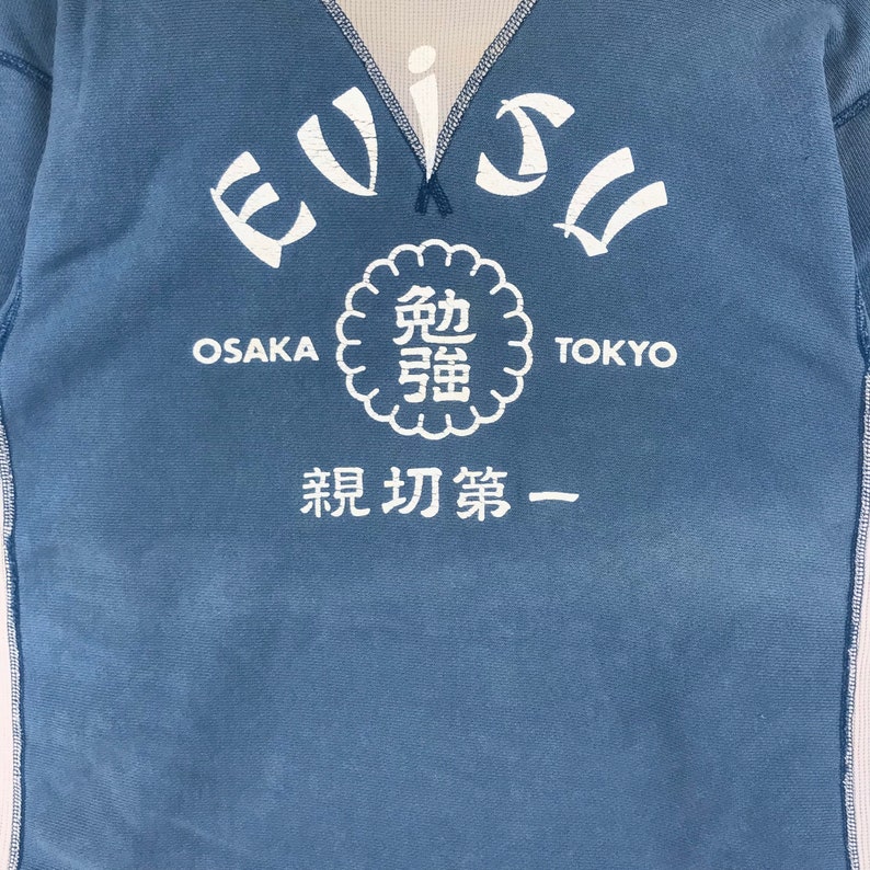 May include: A blue long-sleeved shirt with a white graphic design. The graphic features the text "EVISU" in a curved arc, with the words "OSAKA" and "TOKYO" on either side. A circular graphic with Japanese characters is centred below the text. The text "最初第一" is below the circular graphic.