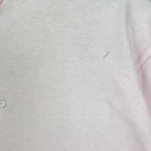 Op de afbeelding: Een lichtroze sweatshirt met een ronde hals. De stof lijkt een zachte brei te zijn. De afbeelding toont een close-up van het bovenste deel van het kledingstuk, met een klein geborduurd detail zichtbaar aan de linkerkant.