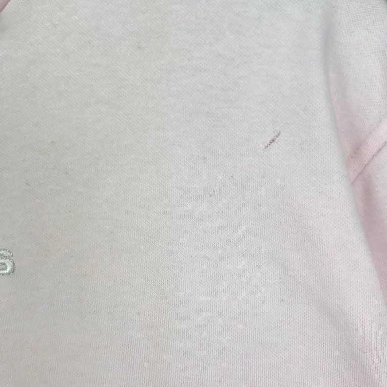 Op de afbeelding: Een lichtroze sweatshirt met een ronde hals. De stof lijkt een zachte brei te zijn. De afbeelding toont een close-up van het bovenste deel van het kledingstuk, met een klein geborduurd detail zichtbaar aan de linkerkant.