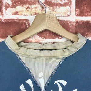 May include: Close-up of the neck of a blue sweatshirt with a tan ribbed collar. The collar has a small black tag with the word "Off" printed on it.