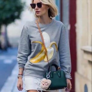 May include: A gray sweatshirt with a yellow banana graphic. The sweatshirt is paired with a white skirt and a small green handbag with a furry keychain.
