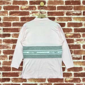 Op de afbeelding: Een lichtroze shirt met lange mouwen, een opstaande kraag en een grijze horizontale streep over de borst. De streep heeft de tekst "courr&egrave;ges sport futur" in het wit. Het shirt hangt aan een houten hanger tegen een bakstenen muur.