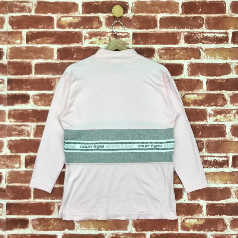 Op de afbeelding: Een lichtroze shirt met lange mouwen, een opstaande kraag en een grijze horizontale streep over de borst. De streep heeft de tekst "courr&egrave;ges sport futur" in het wit. Het shirt hangt aan een houten hanger tegen een bakstenen muur.