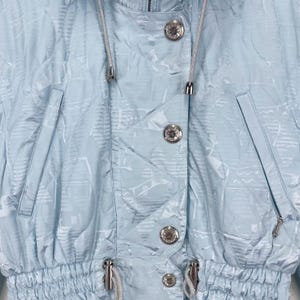 K&ouml;nnte beinhalten: Eine hellblaue Jacke mit einem wei&szlig;en und hellblauen Muster. Die Jacke hat einen Kordelzug in der Taille und eine Kapuze. Die Jacke hat silberne Kn&ouml;pfe und einen Rei&szlig;verschluss.