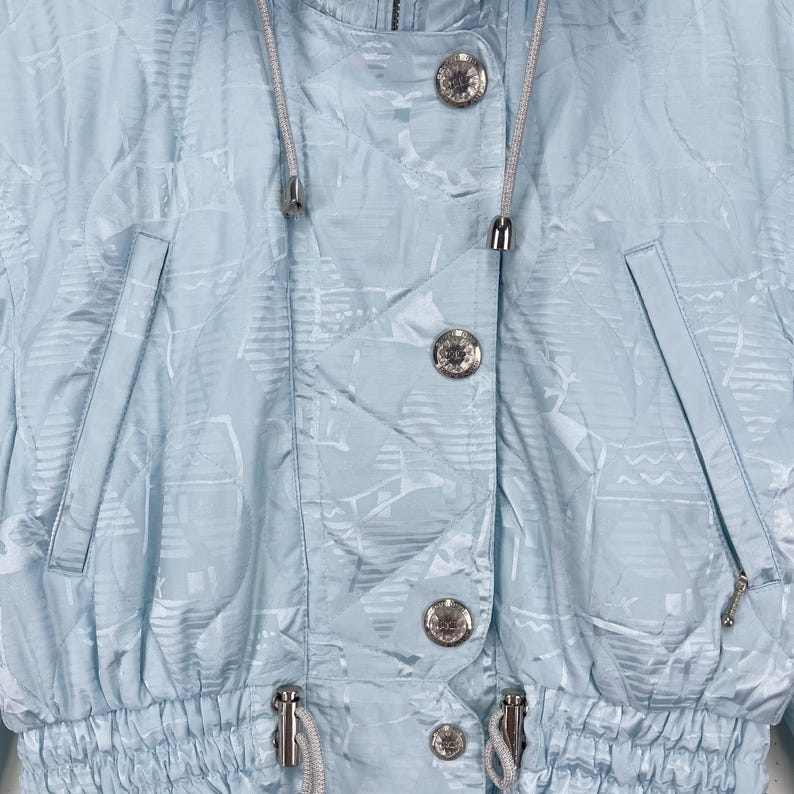 K&ouml;nnte beinhalten: Eine hellblaue Jacke mit einem wei&szlig;en und hellblauen Muster. Die Jacke hat einen Kordelzug in der Taille und eine Kapuze. Die Jacke hat silberne Kn&ouml;pfe und einen Rei&szlig;verschluss.