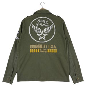 以下が含まれることがあります： オリーブグリーンのミリタリースタイルのジャケット。ボタンダウンフロントと襟付き。背面には、翼、星、そして「Gateway to Success」のテキストが入った白い円形のグラフィックがあります。「DURABILITY U.S.A.」と「33rd BOMB.SQD.」も表示されています。