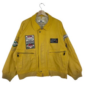 Peut inclure: Veste jaune moutarde avec plusieurs écussons, dont "RACING Mississippi" et "TYPE CWB-IA". La veste a un col, deux poches avant et une fermeture éclair sur la manche gauche. Le texte "ORMANDY" est sur le col.