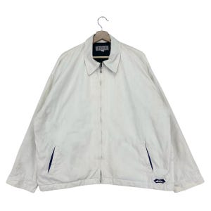 Puede incluir: Una chaqueta blanca con cremallera con un logotipo azul y blanco en la parte inferior derecha. La chaqueta tiene un forro azul alrededor del cuello y los bolsillos.