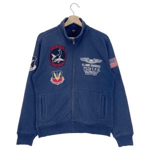 May include: A navy blue zip-up sweatshirt with multiple embroidered patches, including a shark, an American flag, and a military insignia. The text on the patches reads "Uber Shark 75th Fighter Sq", "Flying Sharks 75th T.F.S.  SOVERIGN, MISSION, FORCE, GO, GREAT NAVY", and "1st Combat Command".