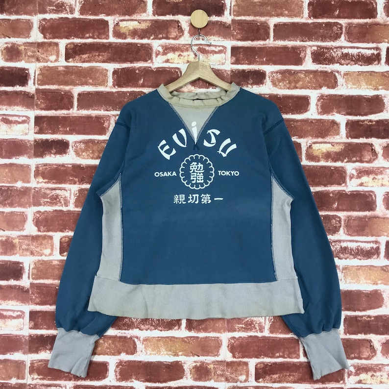 May include: A blue and grey sweatshirt with the text "Evisu" in a white circle with the text "Osaka" and "Tokyo" on either side. The text "鋭刃第一" is below the circle.