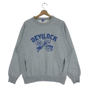 Puede incluir: Sudadera gris con la inscripción azul "DEVILOCK" y un gráfico de antorcha azul. Debajo, se lee "LIGHT MY FIRE". La sudadera tiene cuello redondo y bolsillos laterales, típica de los modelos vintage.