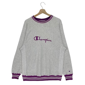 Könnte beinhalten: Ein grauer Champion-Sweatshirt mit violettem Besatz an Hals und Ärmelbündchen. Das Champion-Logo ist in Violett auf der Vorderseite gestickt.