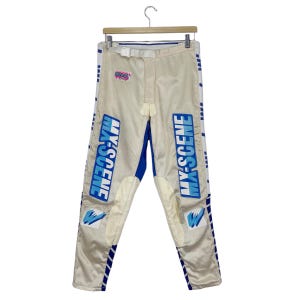 May include: Cream-colored pants with blue and white accents, featuring the text "MX-SCENE" in blue block letters. The pants have a white belt and a small graphic on the upper left. The pants are hanging on a wooden hanger.