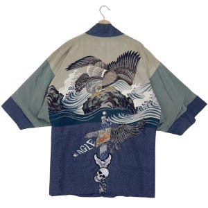 Peut inclure: Un kimono de style japonais bleu et vert avec un motif d'aigle brodé au dos. L'aigle vole au-dessus d'un motif de vagues. Le devant du kimono présente un motif de crâne et d'os croisés avec le mot "EAGLE" écrit au-dessus.