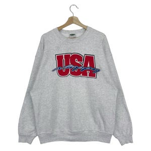 以下が含まれることがあります： グレーのトレーナーに、赤、白、青の「USA」のグラフィックがプリントされています。その下に、青い筆記体で「Happy」と書いてあります。