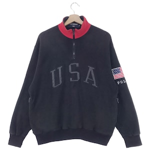 Puede incluir: Sudadera de vellón negra con cremallera roja y un gráfico gris de "USA" en la parte delantera. La manga derecha tiene un pequeño parche de la bandera estadounidense con el texto "POLO" debajo.