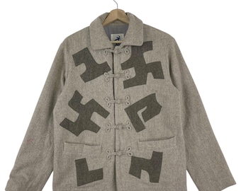 Könnte beinhalten: Beigefarbene Wolljacke mit dunkelgrauen abstrakten Formen. Die Jacke hat einen Kragen, zwei Vordertaschen und Knebelverschlüsse. Die Jacke hängt an einem Holzkleiderbügel.