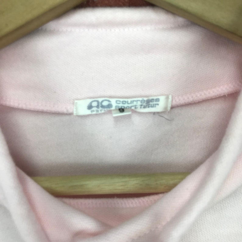 Op de afbeelding: Close-up van een lichtroze poloshirt met een kraag en een label met de tekst "Courr&egrave;ges Sport Tailleur Paris". Het shirt hangt aan een houten hanger.