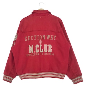 May include: Red wool varsity jacket with a white and beige striped ribbed collar, cuffs, and hem. The back of the jacket features the text "SECTION WAY M.CLUB COLLECTION TO NATURAL" in beige embroidery.