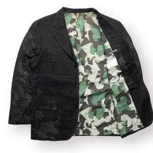 May include: Black blazer with a camouflage pattern on the exterior and a green, black, and white camouflage lining. The blazer has a classic three-button closure and two front pockets. The jacket is open, revealing the patterned interior.