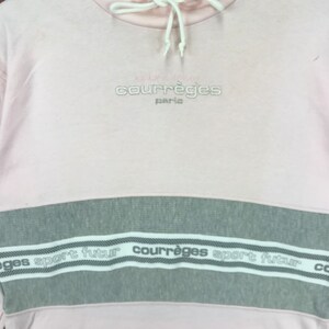 Op de afbeelding: Een lichtroze sweatshirt met een grijze horizontale streep over de borst. Het woord "courr&egrave;ges paris" is in lichtgrijs boven de streep geborduurd. De streep heeft de woorden "sport futur" en "courr&egrave;ges" in wit.